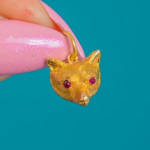 Puede incluir: Un colgante con cabeza de zorro dorado con ojos de piedras preciosas rojas. El colgante se muestra sobre un fondo azul brillante. La cabeza de zorro tiene una textura detallada y un pequeño lazo para colgar.