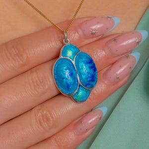 Puede incluir: Un colgante de plata con una cadena dorada. El colgante presenta tres secciones ovaladas de esmalte azul brillante con un borde plateado. El esmalte tiene una calidad iridiscente. El collar se muestra en una persona.