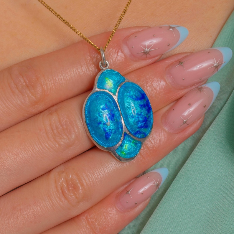 Puede incluir: Un colgante de plata con una cadena dorada. El colgante presenta tres secciones ovaladas de esmalte azul brillante con un borde plateado. El esmalte tiene una calidad iridiscente. El collar se muestra en una persona.