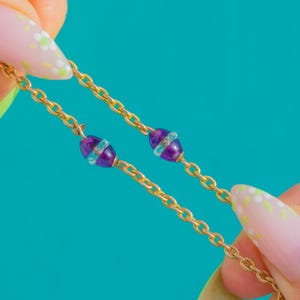 Peut inclure: Un collier en chaîne doré orné de deux perles violettes et bleu clair. Le collier est présenté sur un fond turquoise vif, mettant en valeur les détails du bijou.