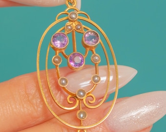 Puede incluir: Colgante dorado con diseño ovalado, con tres piedras preciosas moradas y varias perlas pequeñas. El colgante tiene un marco ornamentado de estilo filigrana con curvas y bucles delicados. El fondo es de color turquesa.