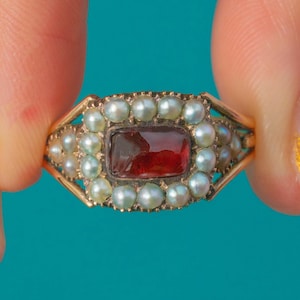 May include: A gold ring featuring a rectangular, deep red gemstone at the center, surrounded by small, round, light-colored pearls. The ring has a delicate, ornate design with a vintage aesthetic.