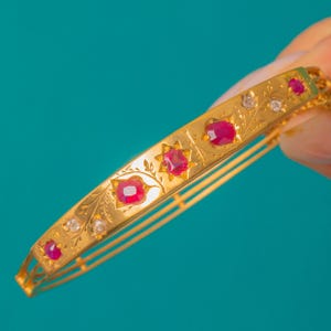 May include: A gold bangle bracelet featuring red gemstones and small clear stones, set within an ornate floral design. The bracelet is displayed against a teal backdrop.
