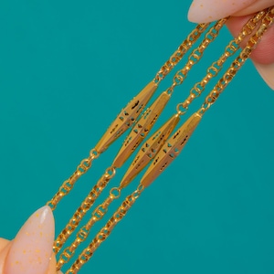 Peut inclure: Gros plan sur un collier en or avec des motifs géométriques complexes. Le collier présente plusieurs rangées de maillons de chaîne délicats et des éléments centraux allongés et ornés. Le bijou est présenté sur un fond turquoise, soulignant sa teinte dorée chaude.
