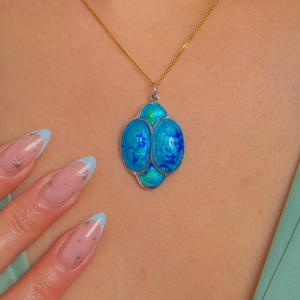 Puede incluir: Un collar con colgante azul y plateado y cadena dorada. El colgante presenta un dise&ntilde;o abstracto con dos &oacute;valos grandes y otro m&aacute;s peque&ntilde;o debajo, en tonos azules y verdes. El collar se lleva alrededor del cuello.