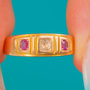 May include: A gold ring featuring three stones set in a horizontal row. The center stone is a clear, round-cut gem, flanked on each side by a smaller, oval-cut vibrant pink gemstone. The band has a smooth, polished finish with vertical metal bars.