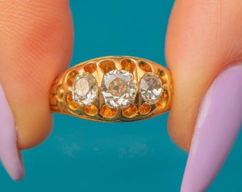 May include: A gold ring with three round diamonds set in a band. The ring is held in a hand with a light purple nail polish.