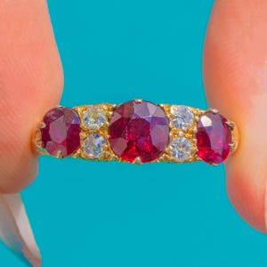 Puede incluir: Un anillo de oro con una gran piedra preciosa de color rojo intenso y corte redondo en el centro, flanqueada por dos piedras rojas más pequeñas a los lados y dos pares de pequeñas piedras blancas transparentes entre ellas.