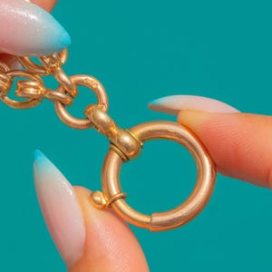 May include: A gold-toned metal spring ring clasp attached to a chain link. The circular clasp features a small protruding lever used to open the mechanism, connected to a series of rounded, interlocking metal links that form a jewelry chain.