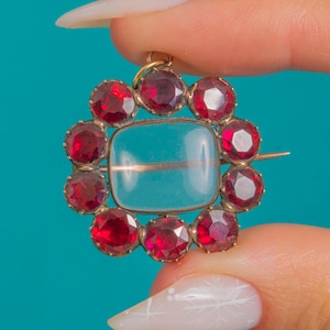 Puede incluir: Broche vintage dorado con una piedra central rectangular azul claro. La broche está rodeada por un círculo de piedras preciosas rojas vibrantes. El alfiler es sostenido por una mano sobre un fondo turquesa.