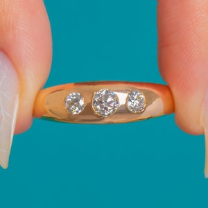 Può includere: Un anello d'oro con tre diamanti rotondi e trasparenti disposti in fila. L'anello è tenuto su uno sfondo turchese brillante, che mette in risalto la fascia in oro lucido e le pietre preziose scintillanti. È un gioiello.