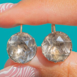 Pode incluir: Um par de brincos de pedra cinza translúcida, redondos e facetados, engastados em armações de metal com ganchos de arame finos e curvos. As pedras apresentam uma superfície multifacetada que reflete a luz, criando um efeito cintilante.