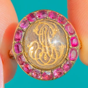 May include: A gold ring with a circular face. The face features a gold monogram inside a clear, circular stone, surrounded by a halo of small, oval-cut pink gemstones. The ring's band is thin and gold.