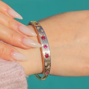 Puede incluir: Brazalete rígido articulado de plata y oro con piedras preciosas rojas y pequeñas piedras transparentes. El brazalete tiene un diseño de estrella y floral. El brazalete está en una muñeca.