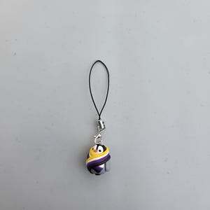 May include: A penguin charm with a black cord and a silver-coloured clasp. The penguin is wrapped in a scarf with the non-binary pride flag colours: yellow, white, purple, and black. The penguin has a black head and a yellow beak.
