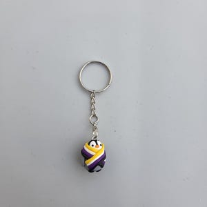 May include: A keychain with a small penguin charm, wrapped in the colours of the non-binary pride flag. The penguin has a black and white body, with a yellow, white, purple, and black striped wrap. The keychain has a silver ring and chain.