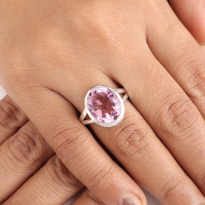 May include: A silver ring featuring a large, oval-cut pink gemstone. The gemstone is set in a simple, elegant band with a split shank design. The ring is shown on a hand.