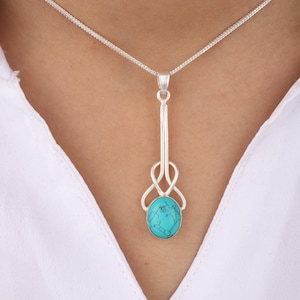 Puede incluir: Un collar con colgante de plata con una piedra ovalada turquesa. El colgante tiene un diseño decorativo con un tallo largo y delgado y un diseño en bucle sobre la piedra. El collar se lleva sobre una camisa blanca.