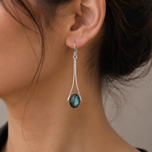 May include: Silver drop earrings with a teardrop design. Each earring features a long, slender silver wire that suspends a dark blue-green, oval-shaped stone. The earrings are worn on a person's ear.
