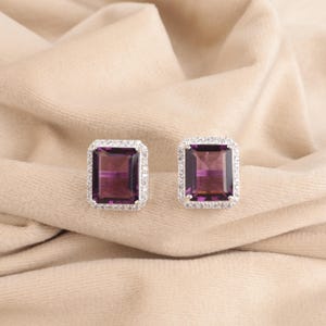 Op de afbeelding: Een paar zilveren oorbellen met een vierkante paarse edelsteen omringd door een halo van kleine diamanten.