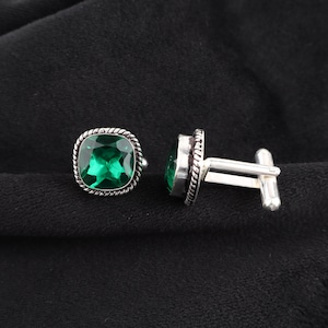 Op de afbeelding: Een paar zilveren manchetknopen met vierkante, smaragdgroene edelstenen. De edelstenen zijn gezet in een zilveren montuur met een gedraaid touwontwerp. De manchetknopen hebben een klassieke T-bar sluiting.