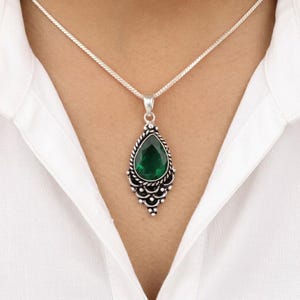 Puede incluir: Un collar de plata con un colgante en forma de lágrima. El colgante presenta una gran piedra preciosa verde esmeralda facetada, engastada en un marco ornamentado de estilo filigrana. El collar se lleva sobre una camisa blanca.