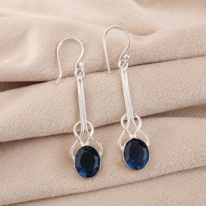 Puede incluir: Un par de pendientes colgantes de plata con piedras preciosas ovaladas de color azul oscuro. Los pendientes tienen un diseño largo y delgado con un detalle de bucle decorativo sobre la piedra preciosa.