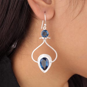 Puede incluir: Pendientes de plata con una piedra preciosa de topacio azul en forma de lágrima y una piedra preciosa de topacio azul redonda más pequeña.