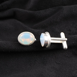 May include: A pair of silver cufflinks with oval-shaped opal stones. The stones are a pale blue color with a hint of pink.