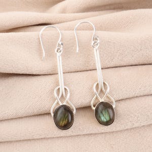 Puede incluir: Un par de pendientes de plata con un diseño largo y delgado. Cada pendiente presenta una piedra preciosa ovalada y oscura con tonos verdes y dorados, engastada en un marco de plata decorativo. Cierre de gancho.