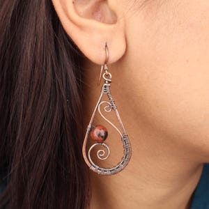 Può includere: Orecchini a goccia in filo di rame con una perla di pietra rossa e marrone. Gli orecchini presentano un design avvolto in filo con un dettaglio a spirale e intrecciato. Il gancio è un design semplice e curvo.