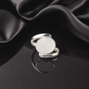 Op de afbeelding: Een zilveren ring met een groot, rond, leeg vlak. De ring heeft een dubbele band, met één band die over het vlak buigt en een andere eronder. De ring wordt getoond op een zwart, reflecterend oppervlak met een zwarte stoffen achtergrond.