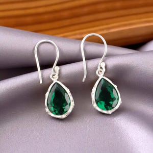 May include: A pair of silver drop earrings featuring teardrop-shaped, emerald-green gemstones. The earrings have a simple design with a hook closure. The gemstones are faceted and set in a silver bezel.