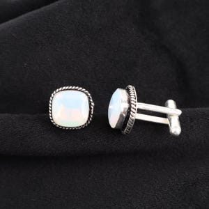 May include: A pair of silver cufflinks with a square, opalescent stone set in a twisted rope-like border. One cufflink is fully visible, while the other is partially visible, showcasing the post and toggle clasp.