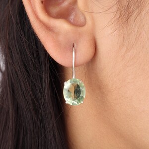 May include: A silver earring with a light green, oval-shaped gemstone. The earring is hanging from the ear, showcasing the gemstone's color and clarity. The design is simple and elegant.