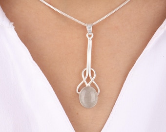 May include: A silver pendant necklace featuring a light gray, oval-shaped stone. The pendant has an intricate, openwork design with a long, slender stem and a chain necklace. The necklace is worn against a white shirt.