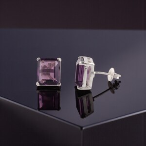 May include: A pair of stud earrings featuring rectangular, light purple gemstones set in silver. The earrings are displayed on a reflective black surface, showcasing their polished appearance and the gemstone's color.