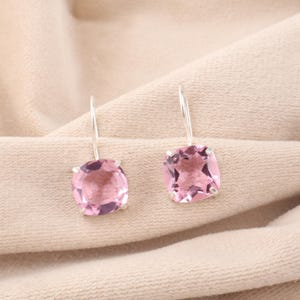Puede incluir: Un par de pendientes colgantes con gemas rosas claras cuadradas engastadas en plata. Los pendientes cuelgan de finos ganchos de plata, mostrando las piedras facetadas. El fondo es una tela beige neutra.