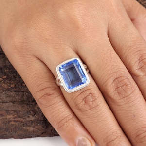 May include: A silver ring featuring a large, rectangular, blue gemstone. The gemstone is set in a silver bezel and has a clear, reflective surface. The ring is displayed on a hand, showcasing its design and color.