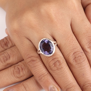 May include: A silver ring featuring a large, oval-cut, deep purple gemstone. The gemstone is set in a simple, polished silver band with a split shank design. The ring is displayed on a hand.