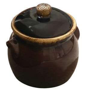 May include: A small, round, dark brown ceramic pot with two side handles and a matching lid. The lid features a glossy black finish with a textured, speckled gold-toned knob at the center. The rim of the pot has a rustic, mottled gold-brown glaze.