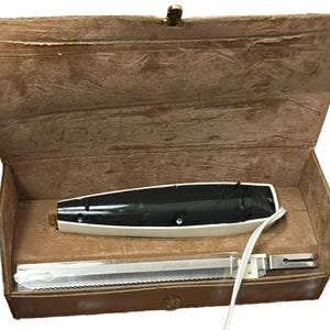 May include: An electric carving knife set in a brown leatherette case. The knife has a white handle with a black top and a stainless steel blade. The case is open, revealing the knife and blade.