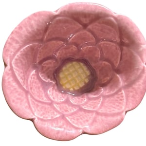 May include: A pink ceramic dish shaped like a flower. The dish has a detailed petal design and a yellow center. The dish is a light pink color and has a glossy finish. The dish is likely used for serving or decoration.