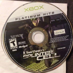 May include: A silver Xbox game disc featuring the game "Tom Clancy's Splinter Cell" with a character graphic. The disc is labeled "Platinum Hits" and includes the ESRB Teen rating and Dolby Digital logo.
