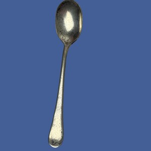 May include: A silver-toned metal spoon with a smooth, oval-shaped bowl and a slender, slightly tapered handle. The surface of the spoon exhibits a weathered, textured patina with visible signs of wear, including dark spots and minor surface imperfections.