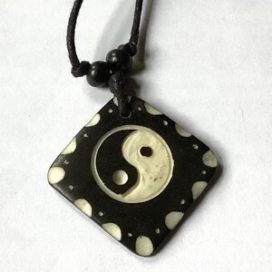 Peut inclure: Un collier en cordon noir avec un pendentif carré représentant un symbole yin yang blanc sur fond noir avec des points blancs.