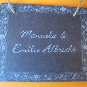 Könnte beinhalten: Ein schwarzes Schiefer-Schild mit einem weißen floralen Rand und dem Text "Manuela & Emilie Albrecht" in weißer Schrift.
