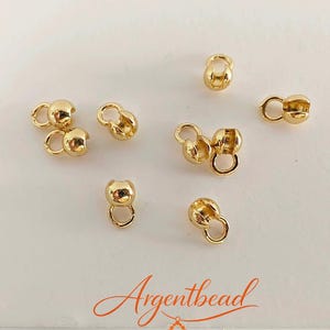 May include: A collection of small, shiny gold-colored crimp cover beads, each featuring a spherical shape with an attached circular loop. The beads are arranged in a scattered pattern, showcasing their metallic finish and functional design.