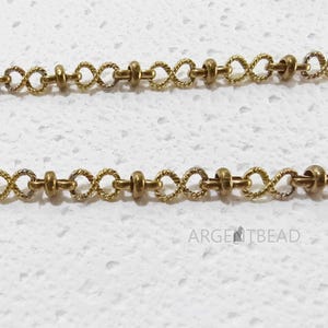 May include: Close-up of two strands of gold-coloured chain. Each chain features a repeating pattern of small, twisted infinity symbols connected by small, round beads. The chains are displayed against a white, textured background. The text "ARGENTBEAD" is visible.