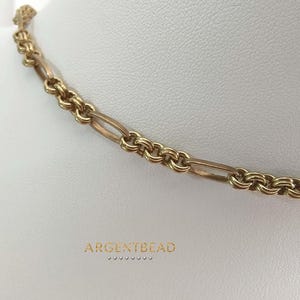 Könnte beinhalten: Eine goldfarbene Kettenhalskette mit einem sich wiederholenden Muster aus länglichen ovalen Gliedern, die durch kleinere, kreisförmige Doppelringsegmente verbunden sind. Der Text ARGENTBEAD steht in goldener Schrift unten.